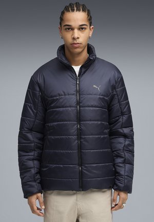 Ung mand med fletninger iført en lynlåset navy blå Puma pufferjakke og beige bukser foran en grå gradient baggrund.