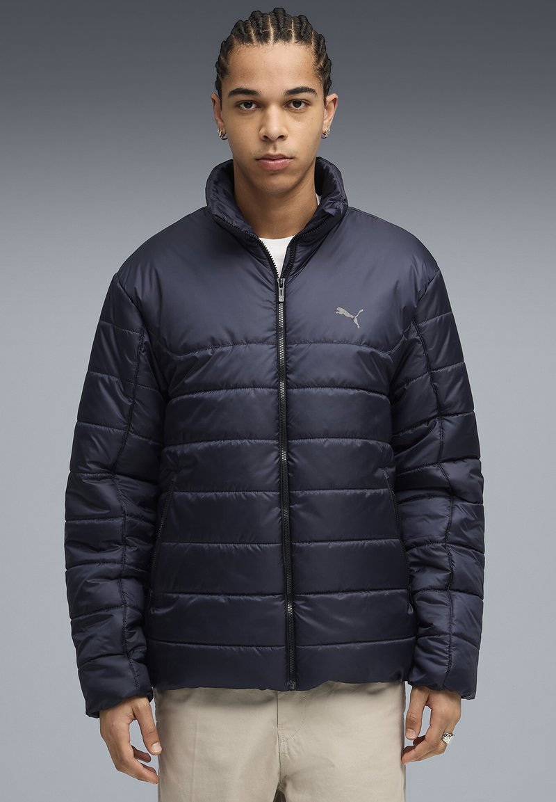 Ung mand med fletninger iført en lynlåset navy blå Puma pufferjakke og beige bukser foran en grå gradient baggrund.