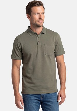 Uomo con capelli castani corti che indossa una polo verde oliva a maniche corte con taschino sul petto e jeans blu, guarda a destra.