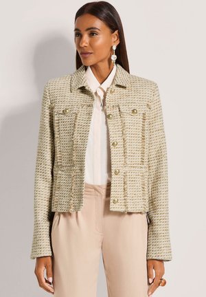 Frau trägt eine beige Tweedjacke mit goldenen Knöpfen über einem weißen Hemd und beigen Hosen, ergänzt durch große hängende Ohrringe.