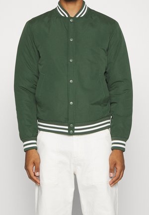 Mann trägt eine grüne Bomberjacke mit weiß gestreiften Bündchen und Kragen, kombiniert mit weißer Hose, vor schlichtem Hintergrund.