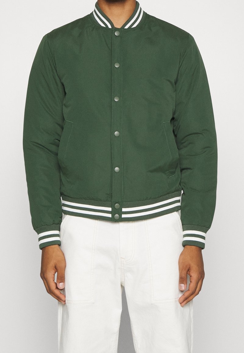 Homme portant un blouson bomber vert avec poignets et col à rayures blanches, associé à un pantalon blanc, debout devant un fond uni.