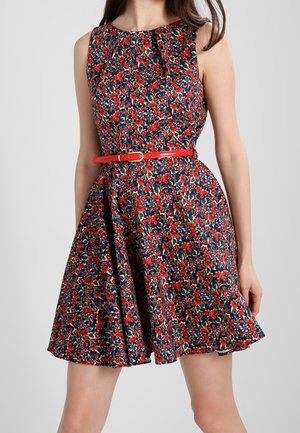 Vrouw draagt een mouwloze donkerblauwe jurk met een rood-wit bloemenpatroon en een smalle rode riem in de taille.