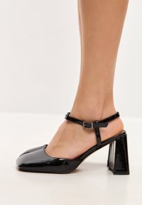 Scarpe con tacco alto in vernice nera, con punta quadrata e cinturino alla caviglia, dotate di un tacco spesso e una texture liscia e lucida.
