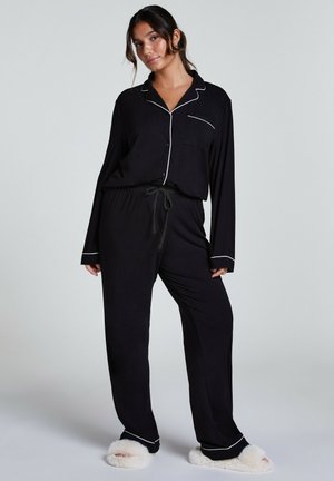 Zwart pyjama set met lange mouwen en broek, voorzien van witte biesdetails. Zachte stof en elastische tailleband voor comfort. Gecombineerd met pluizige slippers.