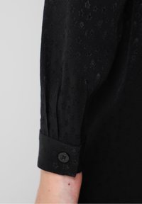 Gros plan d'une manche de chemise noire avec un poignet boutonné, montrant des motifs subtils d'étoiles et de crânes sur le tissu.