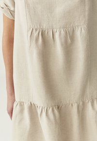 Robe en lin beige avec une taille froncée et un bas évasé. Manches courtes et texture subtile avec des détails de couture visibles.