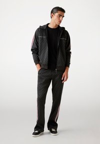Tuta da uomo in tessuto nero a fantasia con strisce laterali rosse e bianche. La giacca presenta un cappuccio e chiusura con zip, mentre i pantaloni sono di taglio ampio.