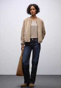 Beige imitationpäls bomberjacka, ljus beige topp, mörkblå jeans, brun mockaväska och matchande skor. Enklare, avslappnad design utan framträdande mönster.