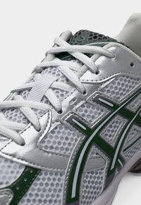 Κοντινή λήψη ενός λευκού αθλητικού παπουτσιού με πλέγμα, ασημί επενδύσεις και πράσινο λογότυπο, με λευκά κορδόνια δεμένα σε διασταυρούμενο μοτίβο.