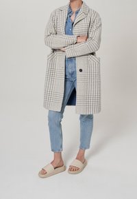 Abrigo de cuadros beige y gris con grandes bolsillos, usado sobre una camisa de mezclilla y combinado con jeans azul claro y sandalias beige.