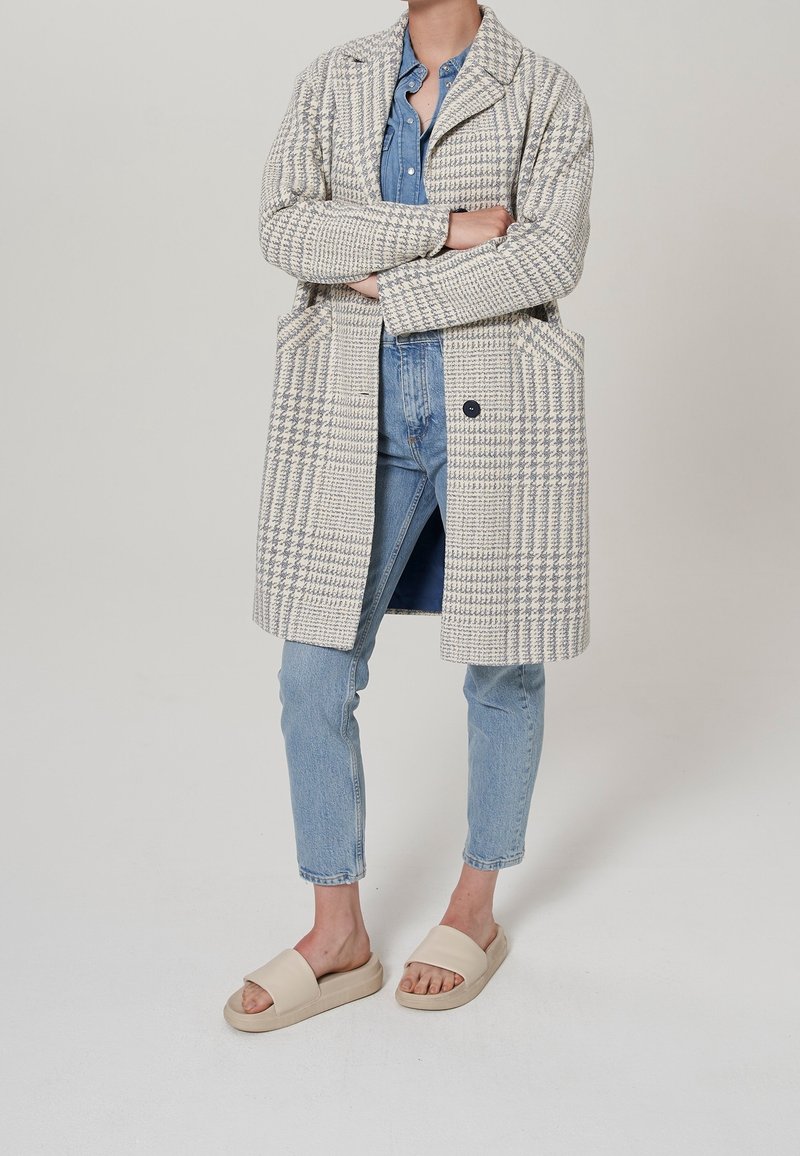 Abrigo de cuadros beige y gris con grandes bolsillos, usado sobre una camisa de mezclilla y combinado con jeans azul claro y sandalias beige.