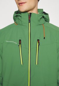 Uomo che indossa una giacca verde Alpine Mountain Wear con cappuccio, cerniere giallo neon sui taschini sul petto e sul davanti, e cordino arancione.
