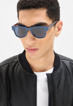 Giovane uomo con occhiali da sole blu, giacca di pelle nera, camicia bianca e un piccolo orecchino a cerchio d'oro su sfondo bianco.