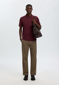 Jeune homme debout, portant un polo à manches courtes bordeaux, un pantalon beige, des chaussures noires et tenant un sac à bandoulière marron foncé.