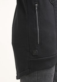 Černá mikina s kapucí s prošívanou texturou, vybavená boční kapsou na zip, značkovou nášivkou a mírně prodlouženým spodním lemem, vyrobená z látky s příměsí bavlny.