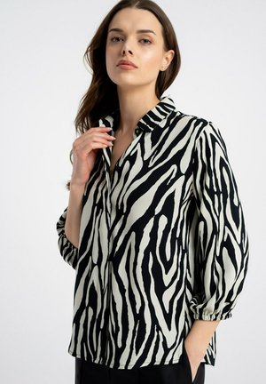 Młoda kobieta z długimi brązowymi włosami ma na sobie czarno-białą bluzkę w zebrę z rękawami 3/4 i kołnierzykiem, pozując na białym tle.