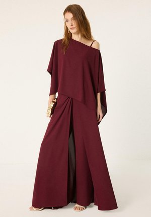 Mujer con vestido burdeos largo hasta el suelo, con hombros descubiertos y una abertura frontal, sosteniendo un clutch dorado y posando frente a un fondo liso.