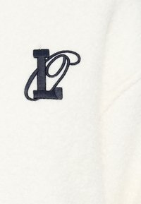 Vêtement en polaire blanche avec un logo 'L' brodé en bleu marine foncé au design fluide. La surface a une apparence douce et texturée.