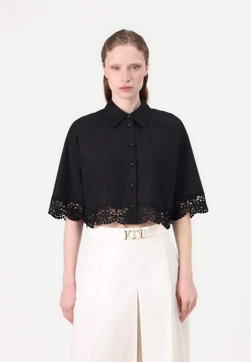 Black cropped blouse with a collar and buttons, featuring intricate lace trim at the hem and a textured finish. Paired with a white high-waisted skirt.
