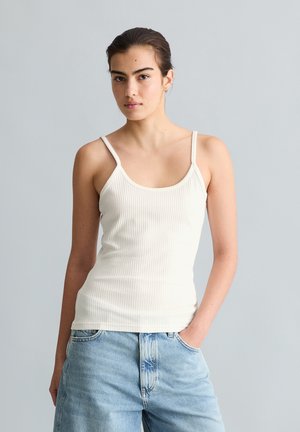 Jeune femme aux cheveux foncés, portant un débardeur blanc côtelé et un jean bleu clair, debout avec une main dans la poche sur un fond gris uni.