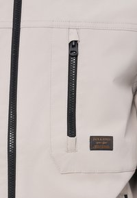 Veste beige avec un tissu texturé, des fermetures éclair noires et un écusson de marque. Poche zippée visible et détails de couture qui améliorent le design.
