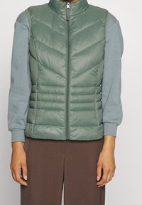 Gewatteerd vest in gedempte groene kleur, met chevronstiksels en een hoge kraag. Gecombineerd met een lichtblauw shirt met lange mouwen en een bruine broek.