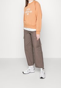 Felpa arancione con logo "new balance" bianco, abbinata a pantaloni cargo marroni e sneakers bianche. L'outfit presenta un design casual.