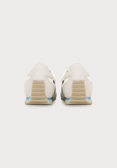 Paire de baskets basses blanches avec talons côtelés beige et accents bleus, vues de dos sur un fond neutre uni.