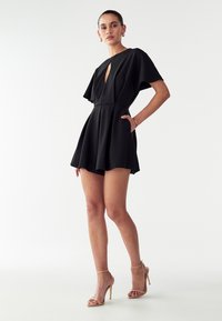 Črna kratka obleka z globokim V-izrezom, letečimi rokavi, prilegajočim pasom in širšim krilom. Ima stranske žepe in gladko teksturo.