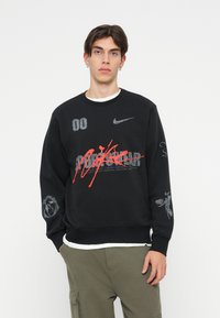 Črna majica s kapuco z grafičnimi potiski, vsebuje Nike logotip, rdeče poudarke in različne vzorce. Ovratnik s krožnim izrezom, rebrasti manšeti in detajl na robu.