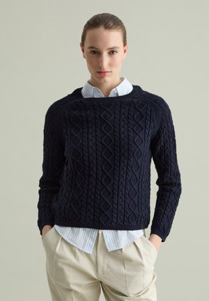Navy knitted sweater with textured diamond patterns, round neckline, and ribbed cuffs, layered over a light blue striped shirt.