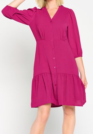 Nainen pukeutuneena fuksianväriseen nappilistaan, jossa on neljänneshihaiset liehukkeet ja röyhelöinen helma, yksinkertaisen taustan edessä.