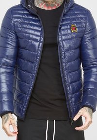 Marinblå vadderad jacka med huva, med horisontell quiltad textur, en dragkedja framtill och en liten broderad logotyp på bröstet.