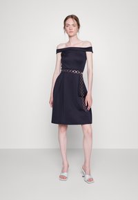 Vestido azul marinho com ombros à mostra, corpo ajustado, saia rodada e um cinto de corrente dourada. O modelo usa sandálias de salto bloco em azul claro.