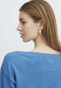 Pull bleu clair à décolleté large et tricot texturé. Des boucles d'oreilles argentées en forme de goutte ajoutent une touche d'élégance. Cheveux coiffés en vagues lâches.