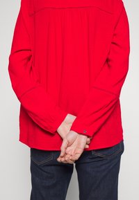 Blouse rouge à manches longues avec un design plissé, featuring des coutures décoratives sur les épaules et des détails de boutons aux poignets, associée à un jean foncé.