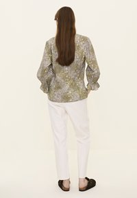 Grön blus med blommönster och volangärmar, kombinerad med vita byxor och mörka slip-on skor. Neutral bakgrund.