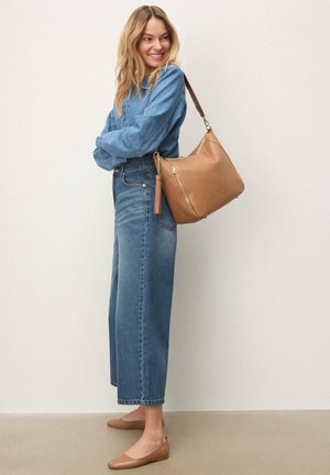 Usměvavá žena v modré džínové košili a džínách stojí bosá a drží tan koženou kabelku přes rameno se zlatým zipem a střapcem na pozadí světlé stěny.