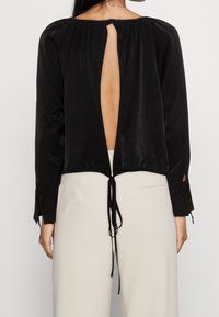 Blouse noire avec un dos ouvert et décolleté plongeant, col froncé et manches longues agrémentées de liens aux poignets. Confectionnée en tissu lisse.