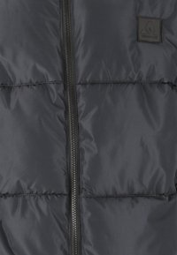 Doudoune noire avec un motif de couture horizontal et une fermeture éclair à l'avant. Présente un patch logo texturé sur le côté supérieur gauche.