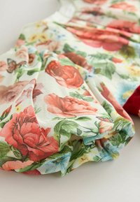Kukallinen mekko kevyestä, teksturoidusta kankaasta, jossa on punaisia, vaaleanpunaisia ja sinisiä kukkia vaaleassa taustassa; sisältää kerrostettuja yksityiskohtia ja punaisen korostuksen.