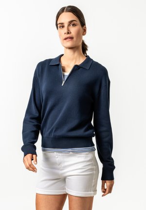 Frau mit dunklem Haar in einem tief gebundenen Pferdeschwanz, die einen marineblauen Pullover mit Reißverschluss am Kragen über einem gestreiften Hemd und weißen Shorts trägt.