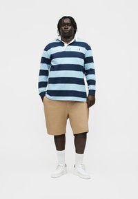 Egy férfi áll, világos és sötétkék csíkos hosszú ujjú políóinget, bézs rövidnadrágot, fehér zoknit és fehér tornacipőt visel, egyszerű háttér előtt.