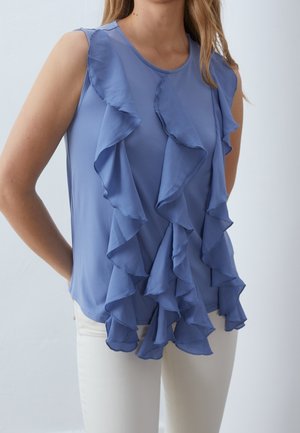 Mouwloze blauwe blouse met gelaagde ruches aan de voorkant, gedragen met witte broek, tegen een effen lichte achtergrond.