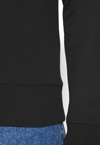 Sudadera negra, de cuello redondo, con dobladillo acanalado, que presenta un tejido texturizado. Combinada con jeans de mezclilla azules para un contraste. Líneas limpias con un detalle mínimo.