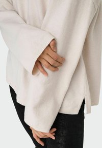 Suéter de punto beige claro con mangas anchas, puños acanalados y aberturas laterales. Textura suave y corte casual, combinado con jeans oscuros.