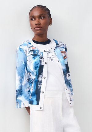 Fonott hajú nő kék-fehér tengerészmintás dzsekiben, fehér ing és fehér nadrág fölött, oldalra néz egy egyszínű háttér előtt.