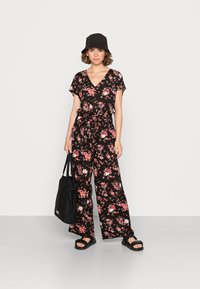 Jumpsuit negro con estampado floral, escote en V, mangas cortas, diseño de pierna ancha, cintura ceñida, combinado con sandalias negras y un bolso tote.