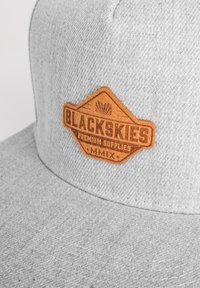 Cappellino grigio con un tessuto testurizzato, con una patch in pelle marrone che presenta "BLACKSKIES PREMIUM SUPPLIES MMIX" impresso sul davanti.
